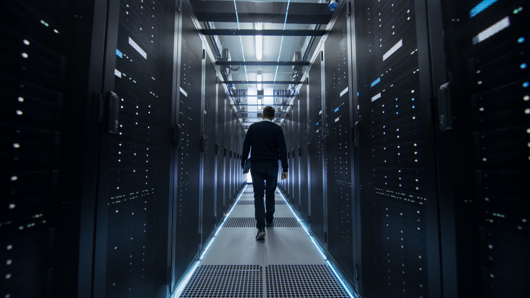 Following Shot of IT Engineer Walking Through Data Center Corridor with Rows of Rack Servers.